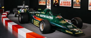 Green Martini Racing Formula 1 car on display in a museum with vintage posters on the wall and red-white barriers in the foreground,