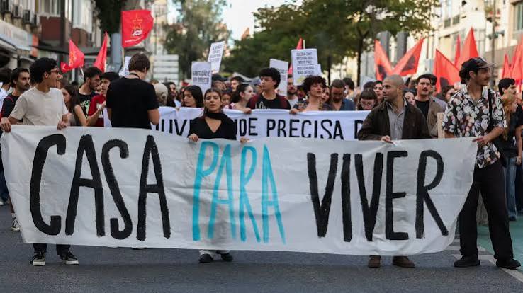 Protest on a city street with a large banner reading 'CASA PARA VIVIR' carried by diverse marchers and red flags in the background.