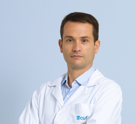 Medical doctor in a white lab coat with arms crossed, facing the camera against a light blue background, CUF logo visible on chest.