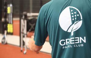 Back of a person wearing a teal Green Padel Club shirt with a white circular logo on the back, on a padel court.