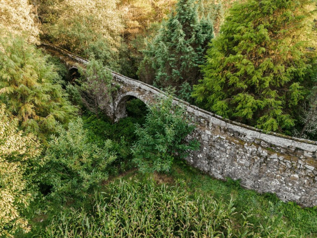 Old stone arch bridge spanning a ravine, surrounded by dense green trees and foliage