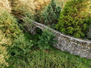Old stone arch bridge spanning a ravine, surrounded by dense green trees and foliage