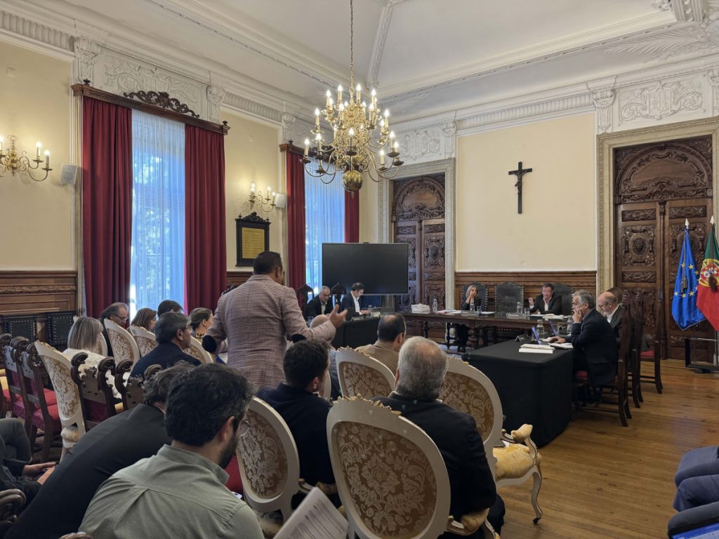 Panel of officials seated at a long table in a richly decorated room with a chandelier, audience, and European/Portuguese flags nearby.