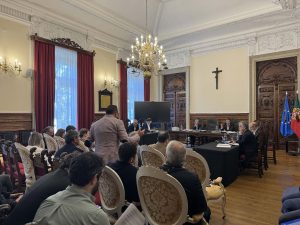 Panel of officials seated at a long table in a richly decorated room with a chandelier, audience, and European/Portuguese flags nearby.