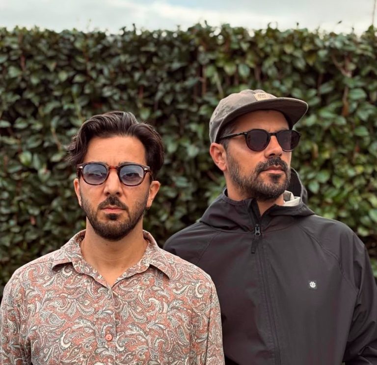 Two men wearing sunglasses stand side by side in front of a leafy hedge outdoors.