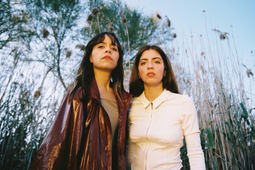 Two women standing side by side in a field of tall reeds, looking at the camera with confident expressions.