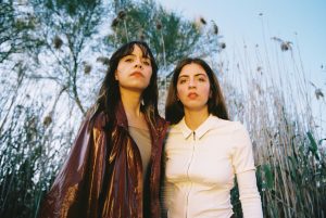 Two women standing side by side in a field of tall reeds, looking at the camera with confident expressions.