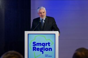 Male speaker in a suit delivering a talk at a podium with a 'Smart Region Summit' banner and blue backdrop.