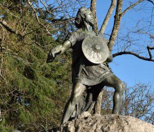 Bronze statue of a warrior with a round shield, sword drawn, standing on a rocky pedestal in a park with leafless trees and a blue sky.