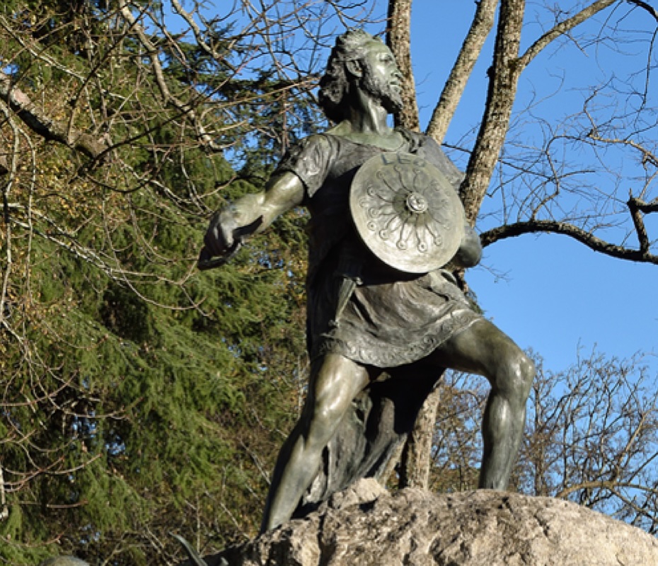 Bronze statue of a warrior with a round shield, sword drawn, standing on a rocky pedestal in a park with leafless trees and a blue sky.